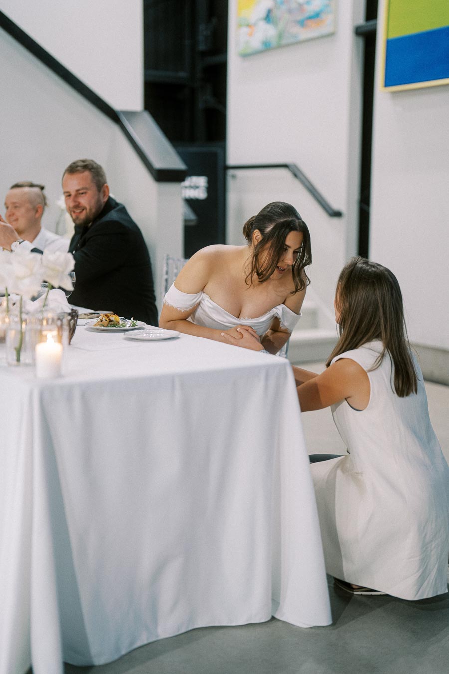 Bride in elegant off-shoulder white gown engaged in conversation at a wedding reception, with other guests seated at a beautifully decorated table with candles and flowers.