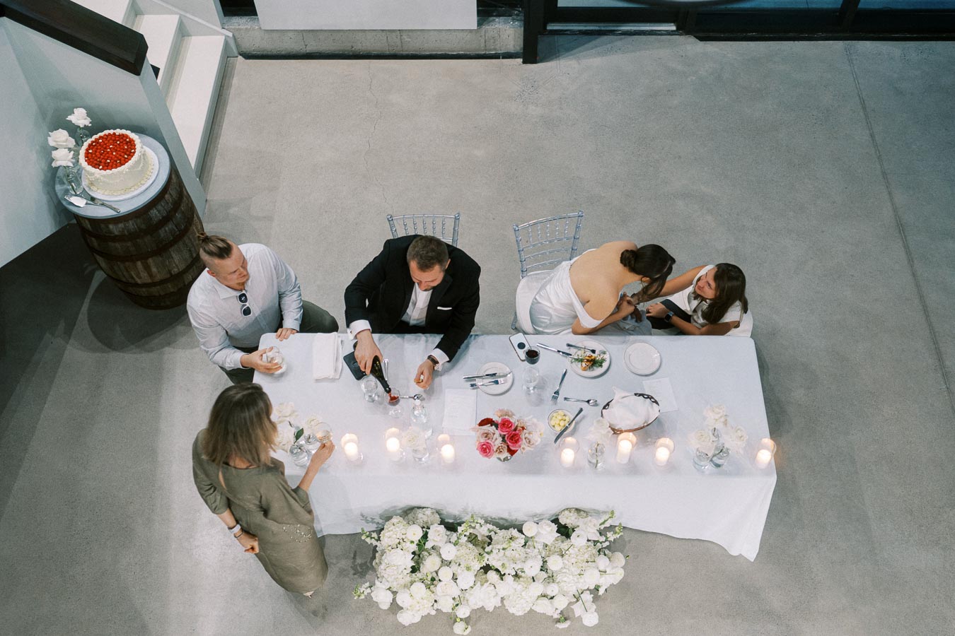 Aerial view of a formal dinner setting with people seated at a white-clothed table, featuring lit candles, elegant flower arrangements, and a two-tiered cake on a barrel stand, creating a sophisticated and celebratory atmosphere.