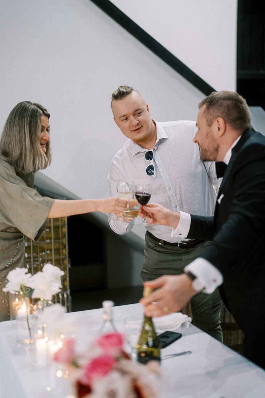 A group of three friends toasting with wine glasses at a celebration, standing around a table decorated with flowers and candles.