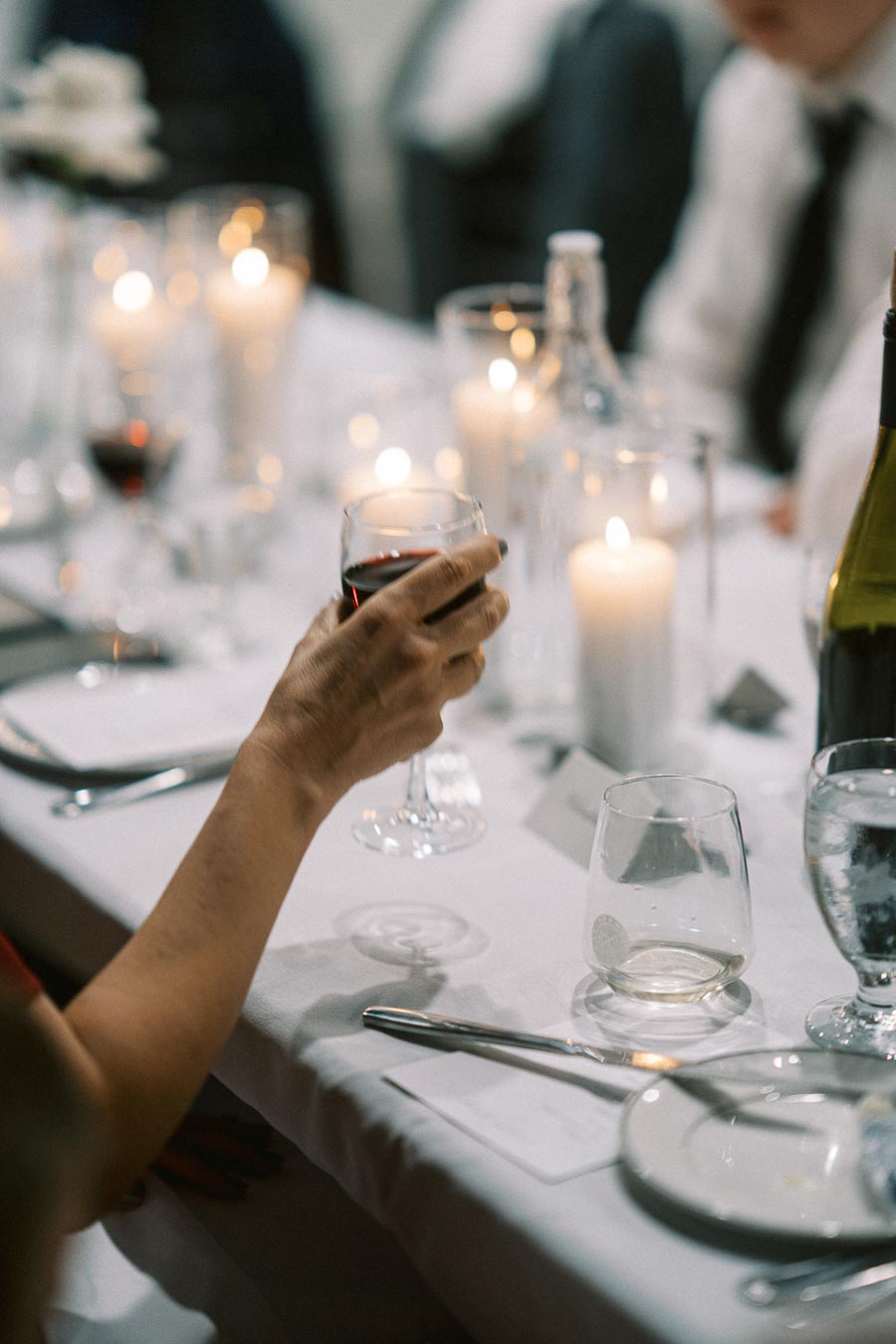 A close-up of an elegantly set dining table featuring a hand holding a glass of red wine, surrounded by lit candles, plates, and glassware, creating a warm and inviting ambiance.