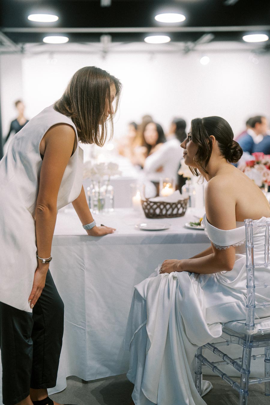 Bride in an elegant white wedding dress sitting and speaking with a wedding planner indoors, surrounded by guests and floral decorations.