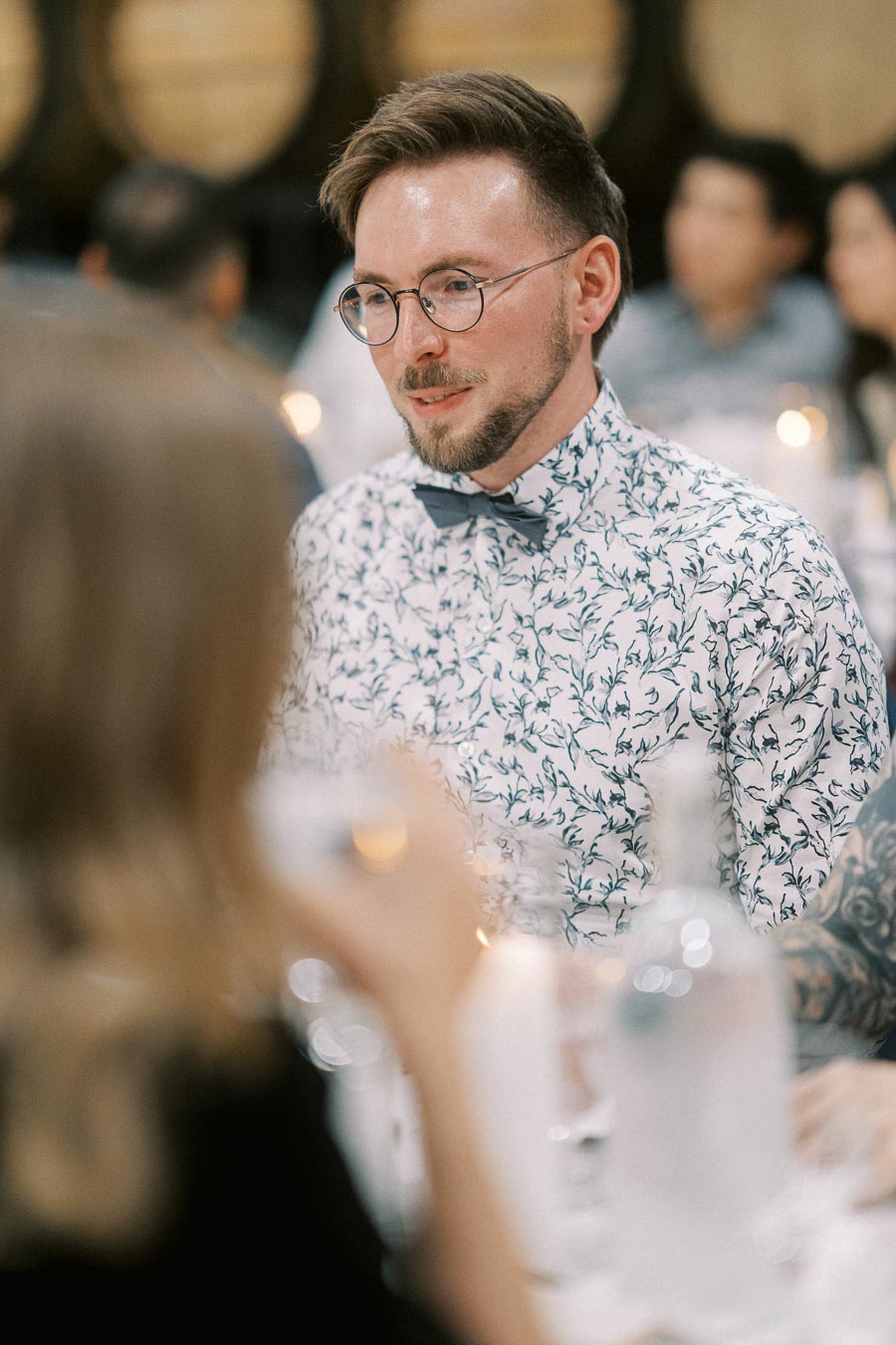 A well-dressed man with glasses and a floral shirt engaging in conversation at a formal event, surrounded by blurred attendees and soft lighting.