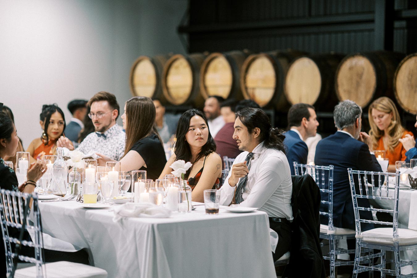 A diverse group of people seated at elegantly decorated tables during a formal dinner event in a rustic setting with wine barrels in the background.