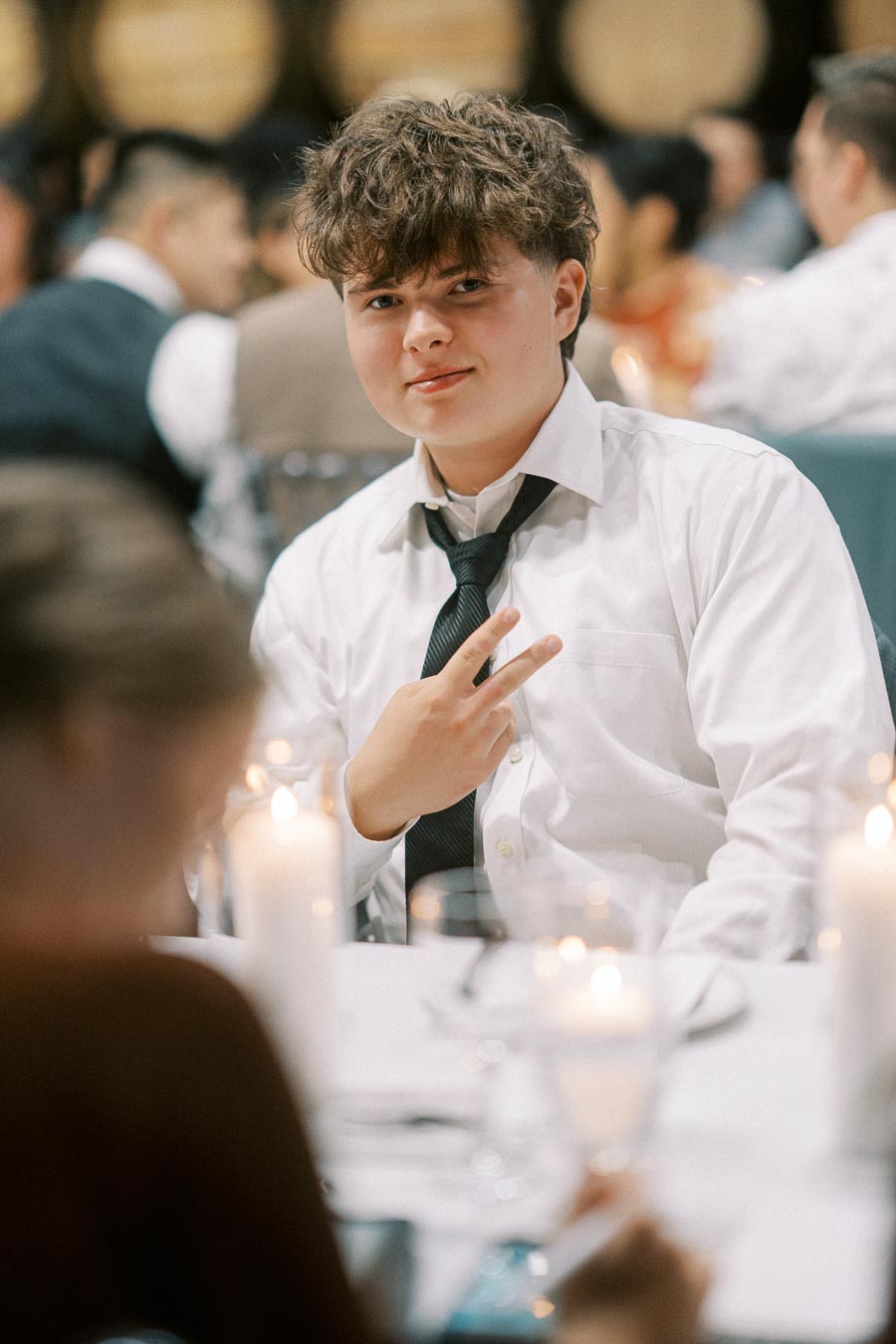 A young person in a white dress shirt and black tie sitting at a candlelit table during an event, making a peace sign gesture amid a blurred background of other guests.