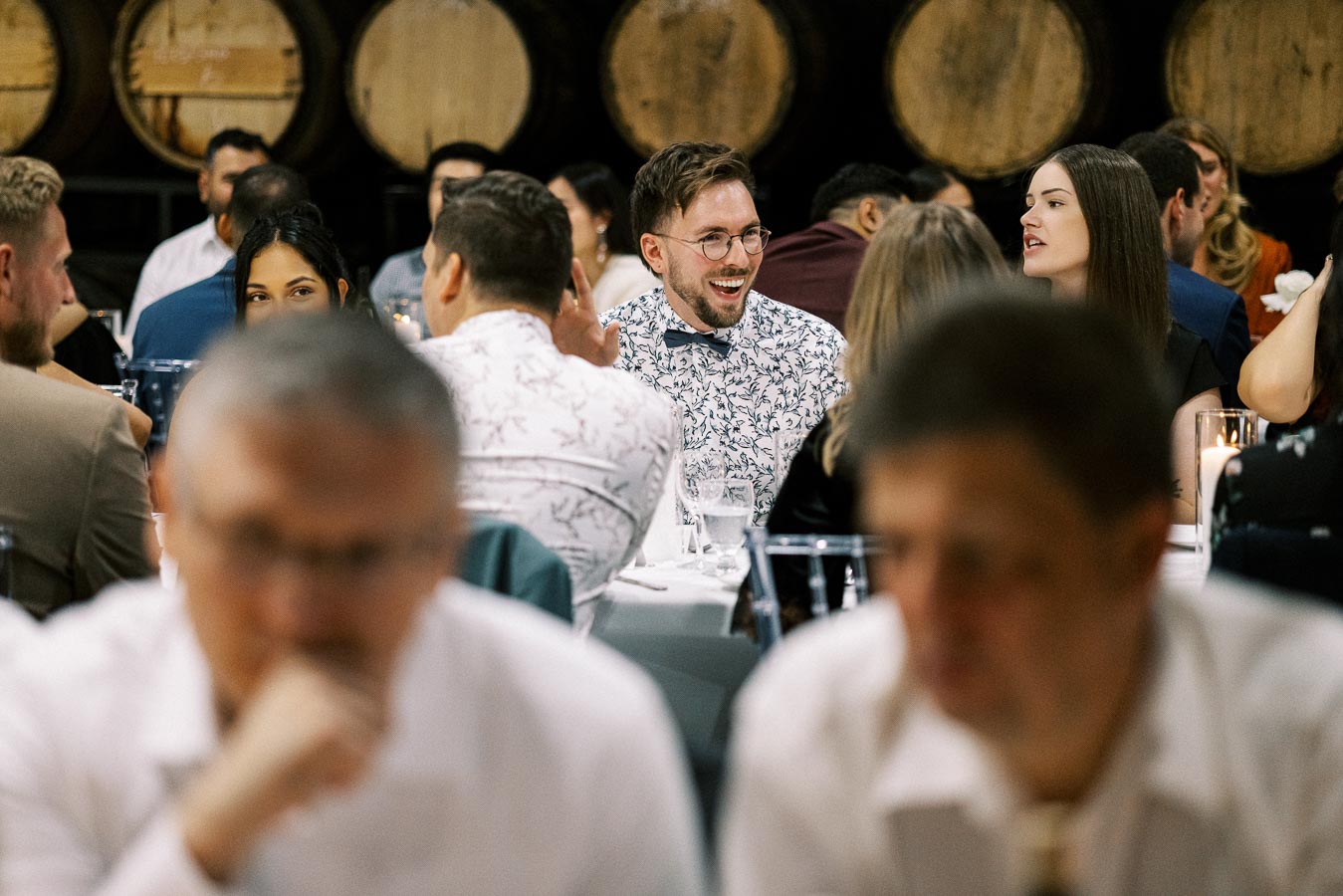 A lively group of people enjoying a social gathering with tables set for dining, surrounded by rustic wooden barrels in a warm, inviting atmosphere.
