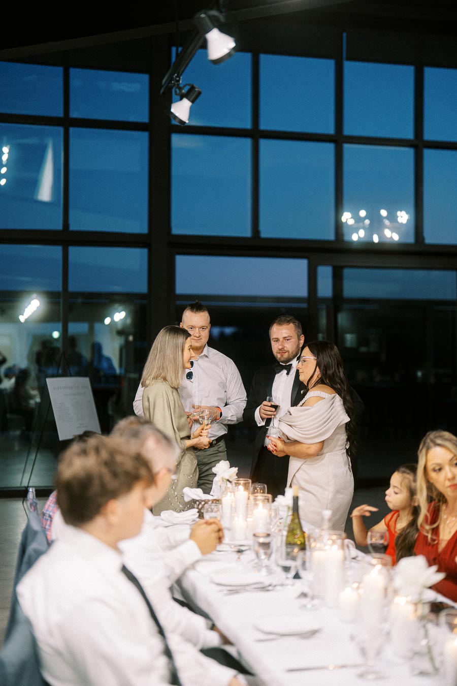 A group of elegantly dressed people socializing around a beautifully set dinner table at a formal evening event inside a modern venue with large windows, candles, and a nighttime backdrop.