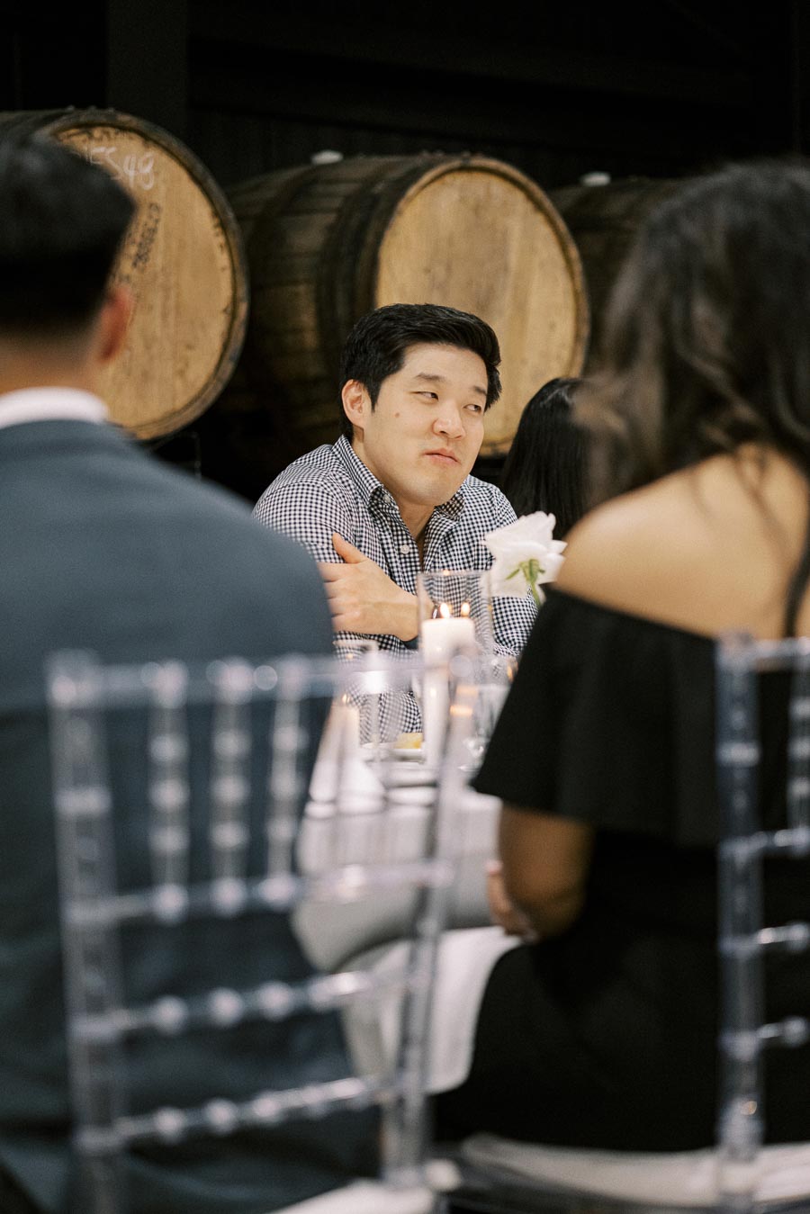 Person in a checkered shirt seated at a dining table in a winery setting, surrounded by candlelight and barrels, engaged in conversation.