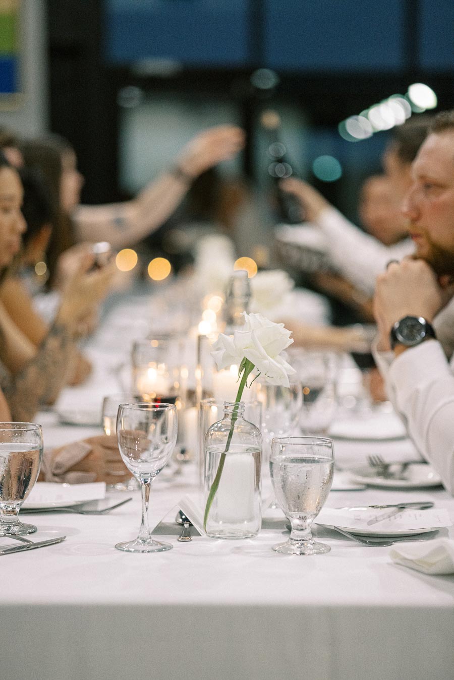 A beautifully set dinner table at a formal event, featuring a white rose in a glass vase, elegant glassware, and candlelight ambiance, surrounded by guests in conversation.