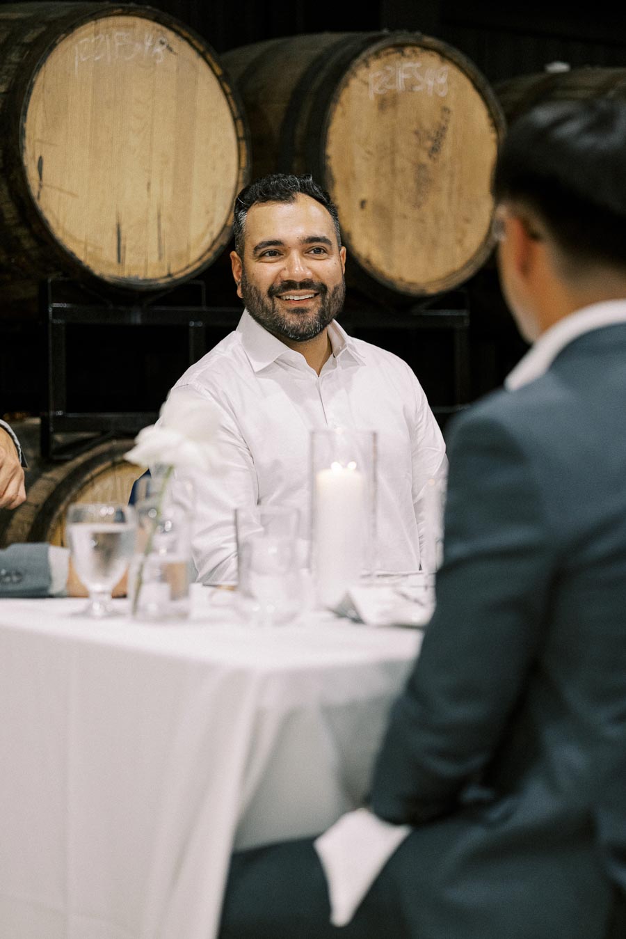 A man smiling and sitting at a white-clothed table in a wine cellar, with large wooden barrels in the background, interacting with others at a formal event.