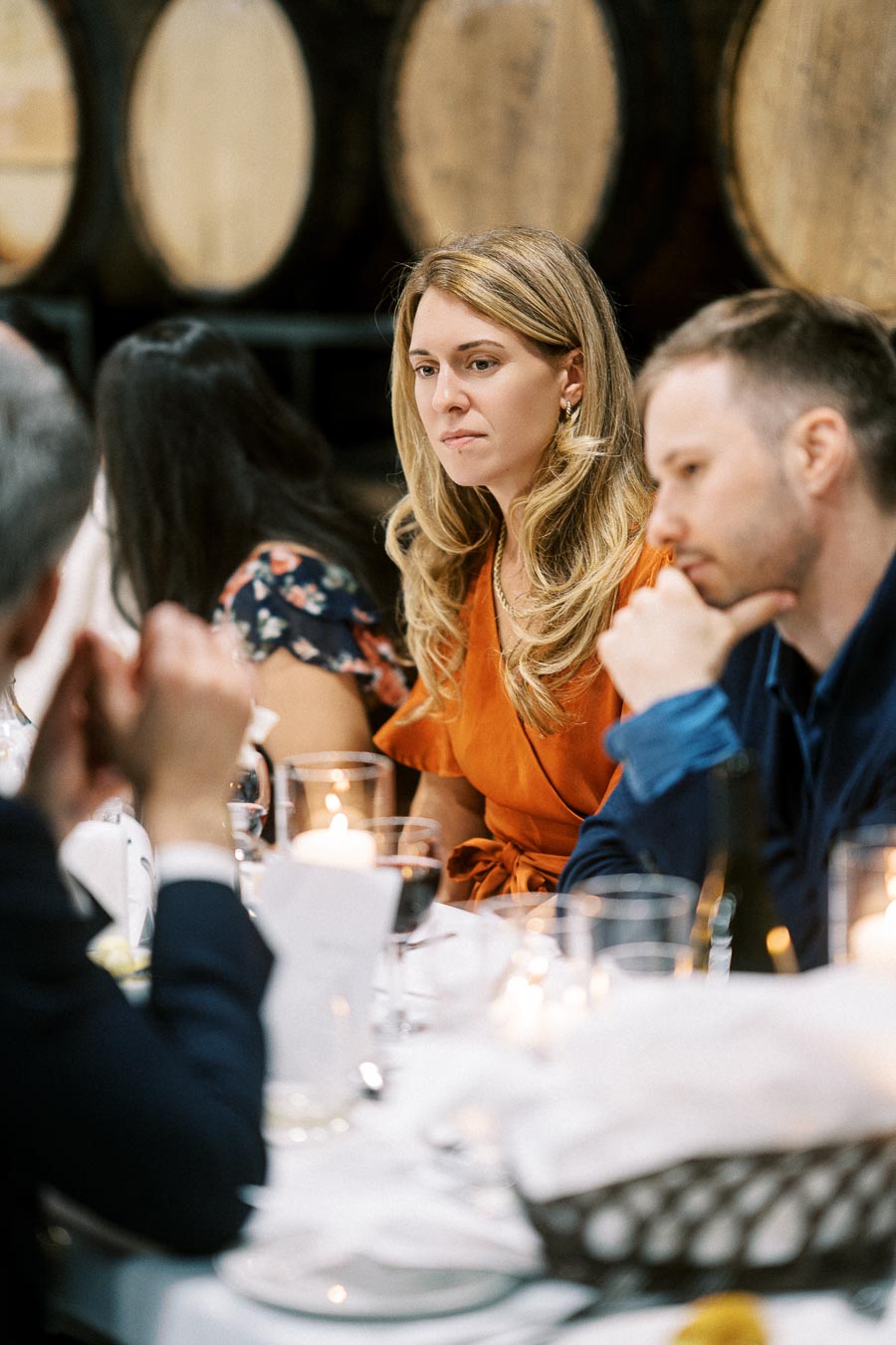 Dinner gathering in a rustic dining setting with a woman in a bright orange dress engaging in conversation, surrounded by other attendees and ambient candlelight.