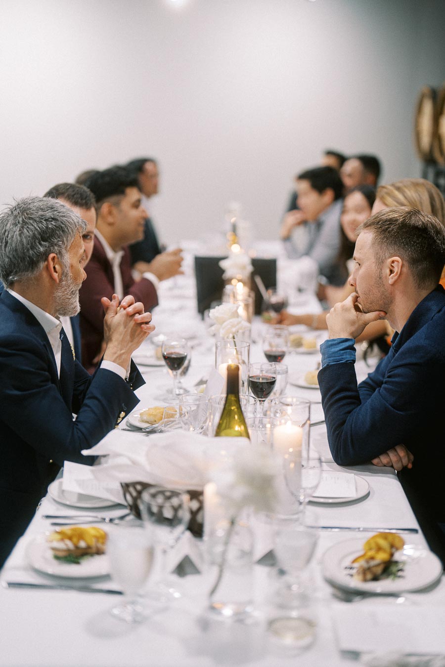 Elegant dinner party setting with a long table, featuring formally dressed guests in conversation, white floral centerpieces, and candlelight ambiance. Plates with gourmet dishes and wine glasses are arranged neatly.