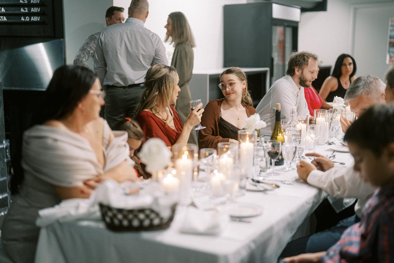 A warm and inviting dinner party scene with people seated around a long, elegantly set table adorned with white flowers and candlelight, engaging in conversation and enjoying drinks.