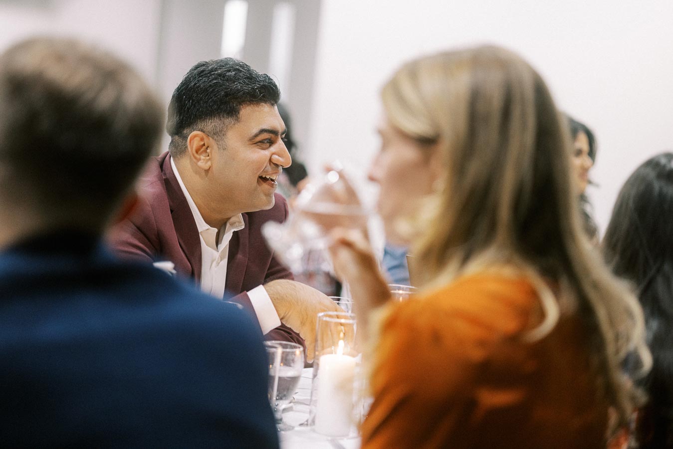 A group of people enjoying a lively conversation at a dinner event, with a man in a burgundy blazer smiling and interacting at a table set with candles and glasses.