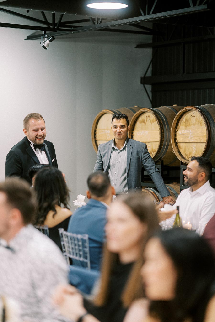 A group of people enjoying a social gathering in a winery setting, with wine barrels in the background and two men in formal attire leading the conversation.