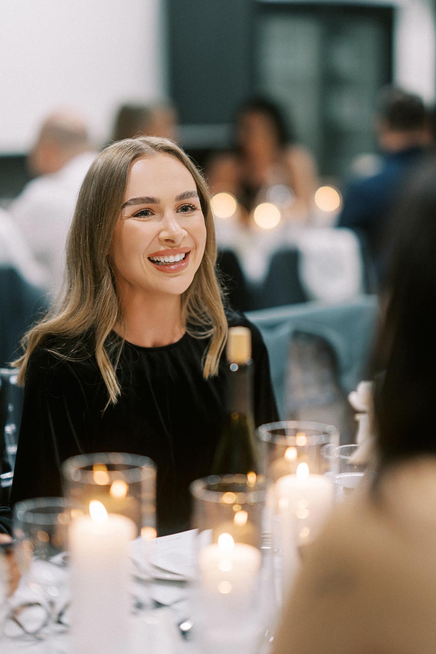A person sitting at a candlelit dinner table, smiling warmly with blurred figures in the background, creating an elegant and festive atmosphere.