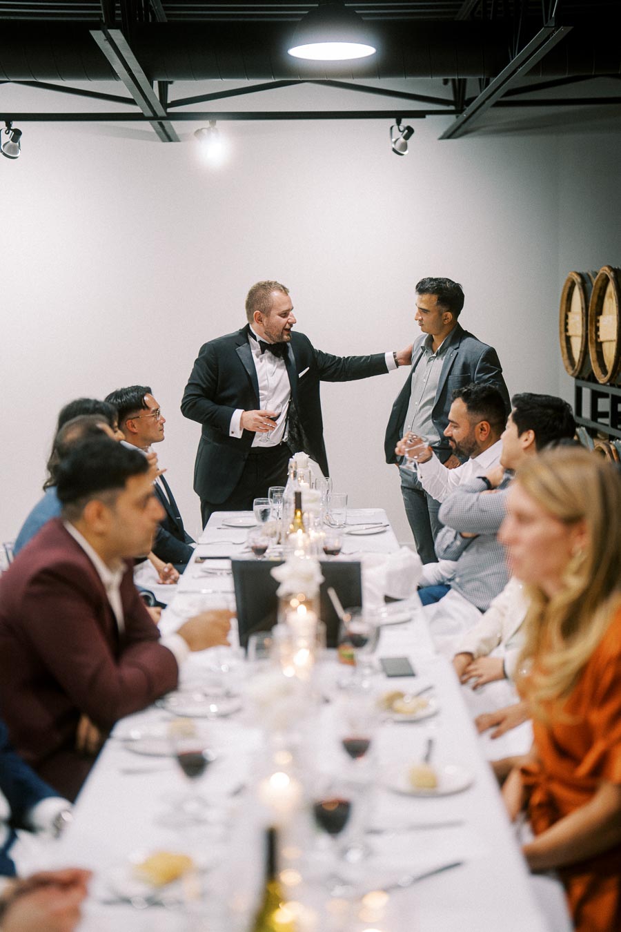 A group of people in formal attire gathered around a dinner table, engaged in conversation at a sophisticated indoor event, with wine glasses and candles creating an elegant atmosphere.