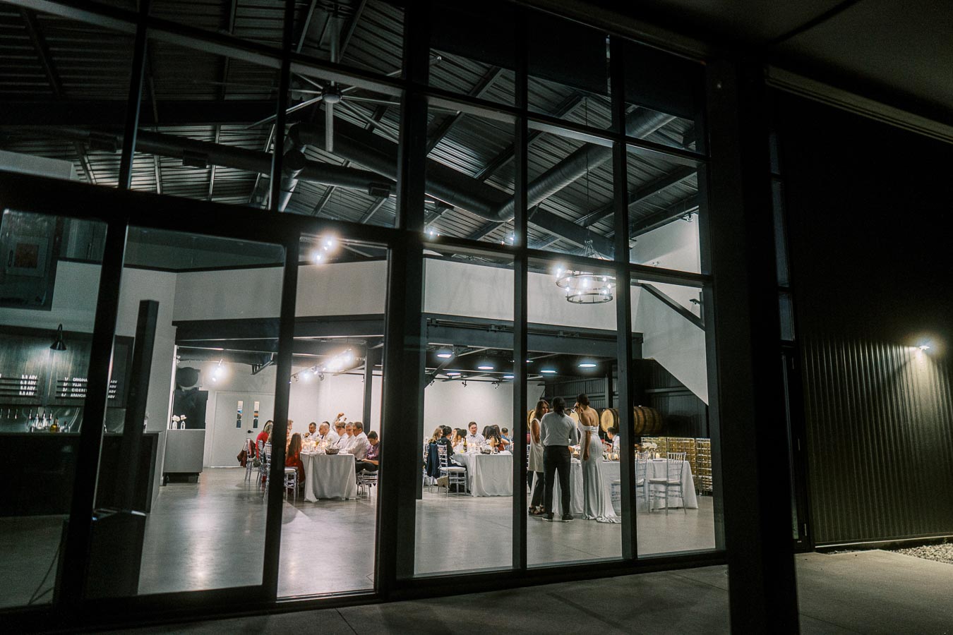 Indoor wedding reception viewed through large glass windows at night, guests seated at decorated tables with ambient lighting, modern industrial venue setting.