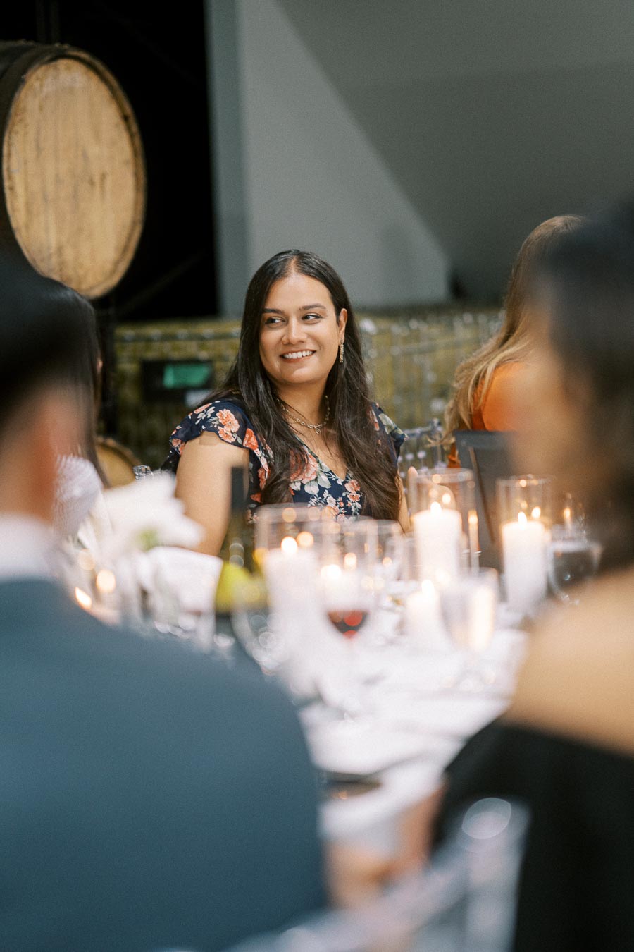 A woman in a floral dress smiling and seated at a candlelit dinner table with people in the background, evoking a warm and festive atmosphere.