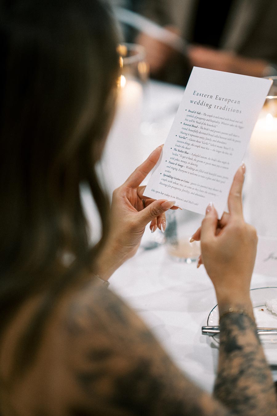 Person reading a card about Eastern European wedding traditions at an elegantly set table, with blurred candlelight ambiance.