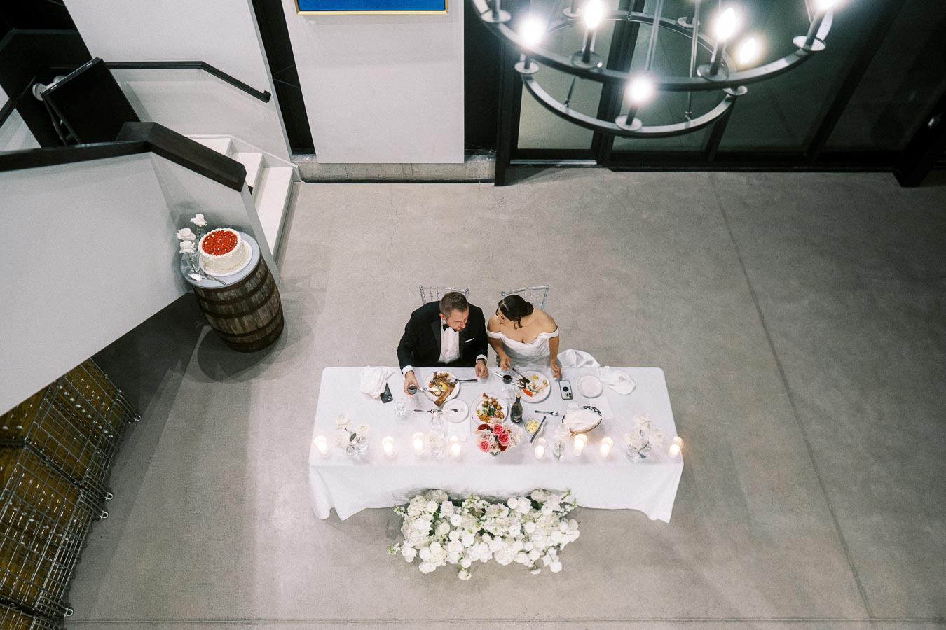 Overhead view of a bride and groom seated at a white-clothed wedding reception table adorned with candles, flowers, and a variety of dishes, in an elegantly decorated venue.