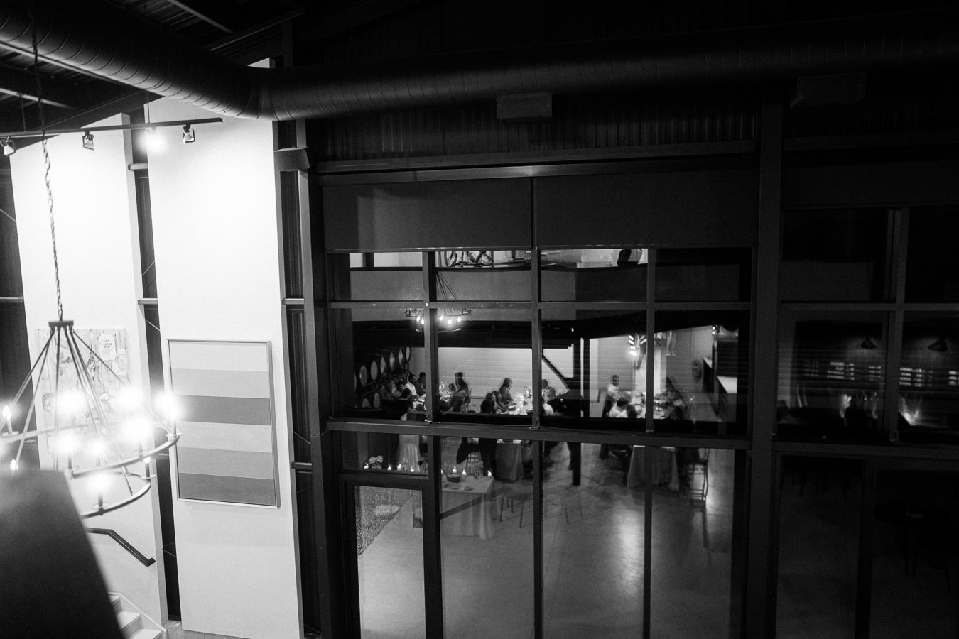 Black and white photograph of an elegant restaurant interior viewed from a high angle, showcasing guests dining at tables through large glass windows, with modern light fixtures and decorative art on the walls.