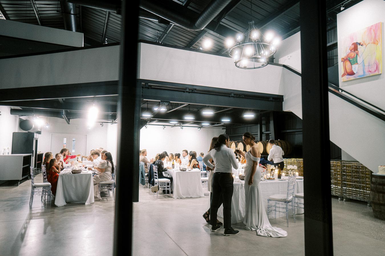 A wedding reception in a stylish industrial venue, featuring guests seated at elegantly decorated tables with white tablecloths, as a couple converses near a circular bar in the background, under modern chandeliers and surrounded by exposed beams and barrels.