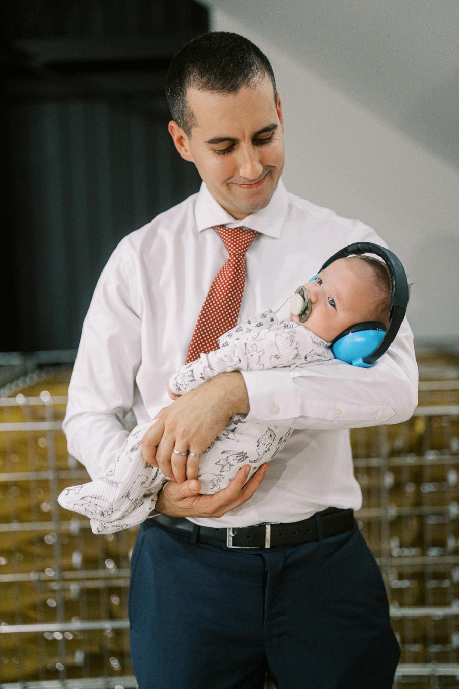 A smiling man in a white shirt and red tie, holding a baby wearing noise-canceling headphones and a pacifier, inside an industrial setting.