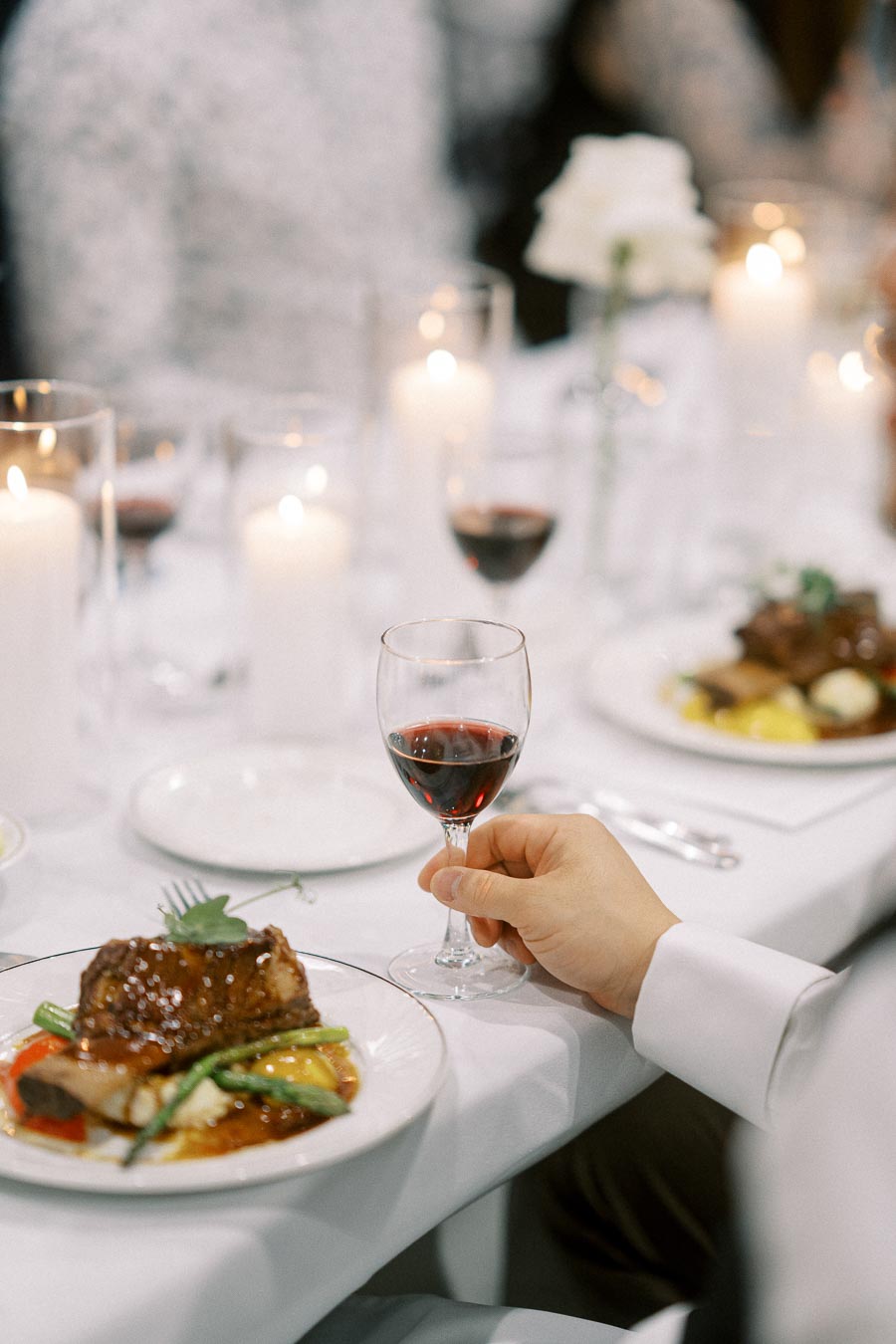A hand holding a glass of red wine at a formal dinner table set with white candles and plates of gourmet food, suggesting an elegant dining experience.