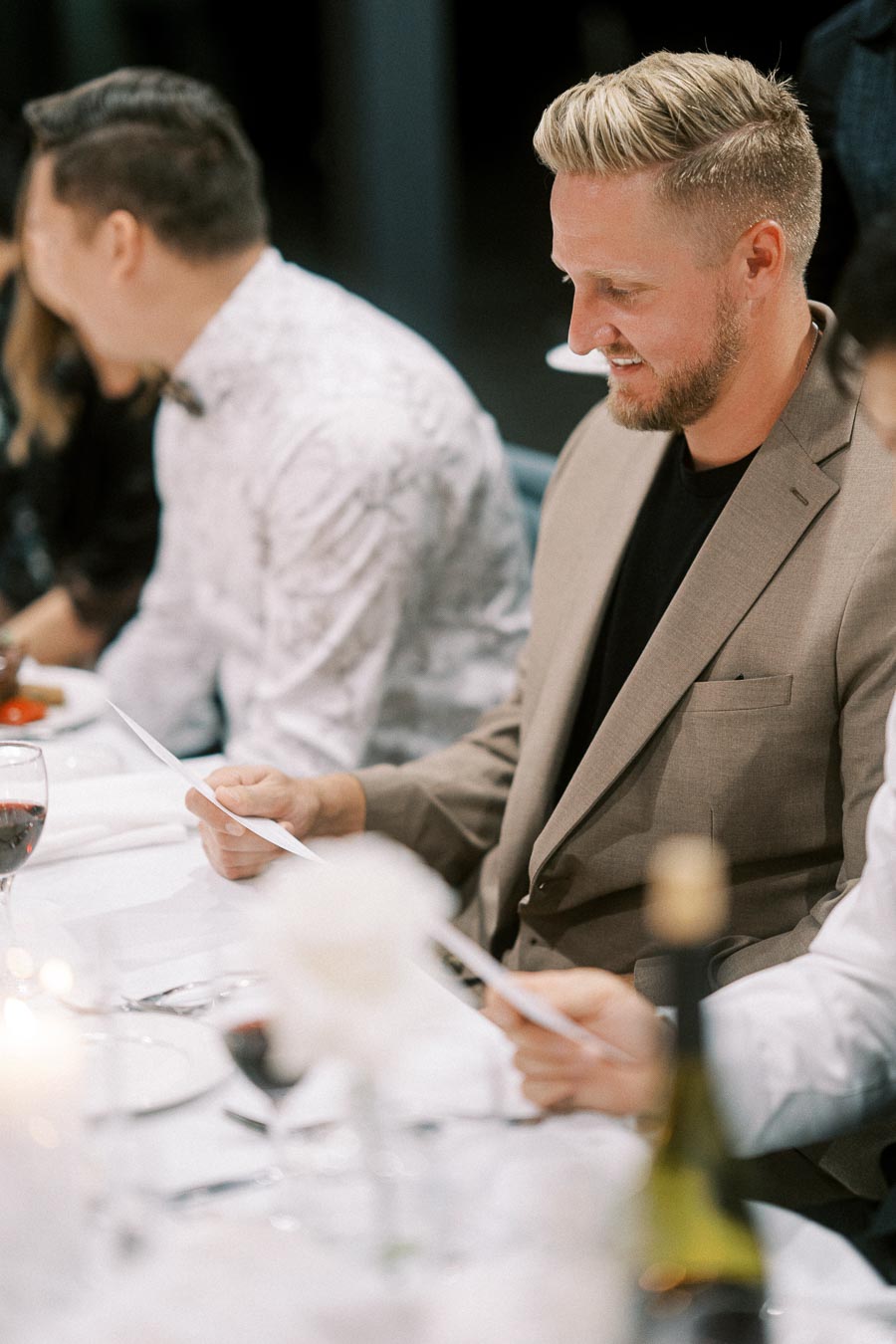 Guests reviewing menus at an elegant dinner event, featuring a well-dressed man with a beige blazer seated at a white tablecloth setting. Wine glasses and a bottle are visible.