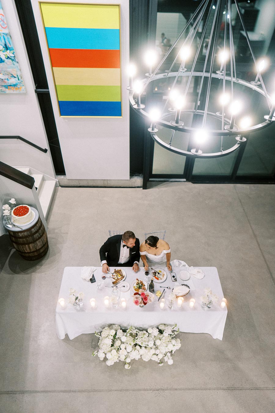 Overhead view of a couple in formal attire dining at a decorated table for a wedding reception, surrounded by elegant floral arrangements and warm candlelight, with modern art and chandelier in the background.