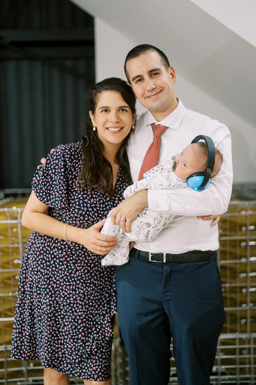 A smiling couple standing together while the man holds a baby wearing blue earmuffs, indoors, with a cozy atmosphere.