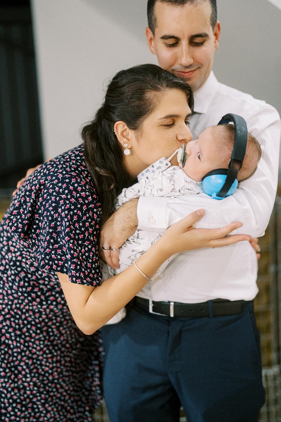 A loving couple holding their baby wearing blue noise-canceling headphones, with the mother giving the baby a gentle kiss.