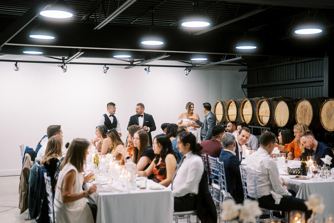 Wedding reception in a modern winery setting with guests seated at elegantly decorated tables, large wine barrels in the background, and a newlywed couple standing nearby engaging with guests.