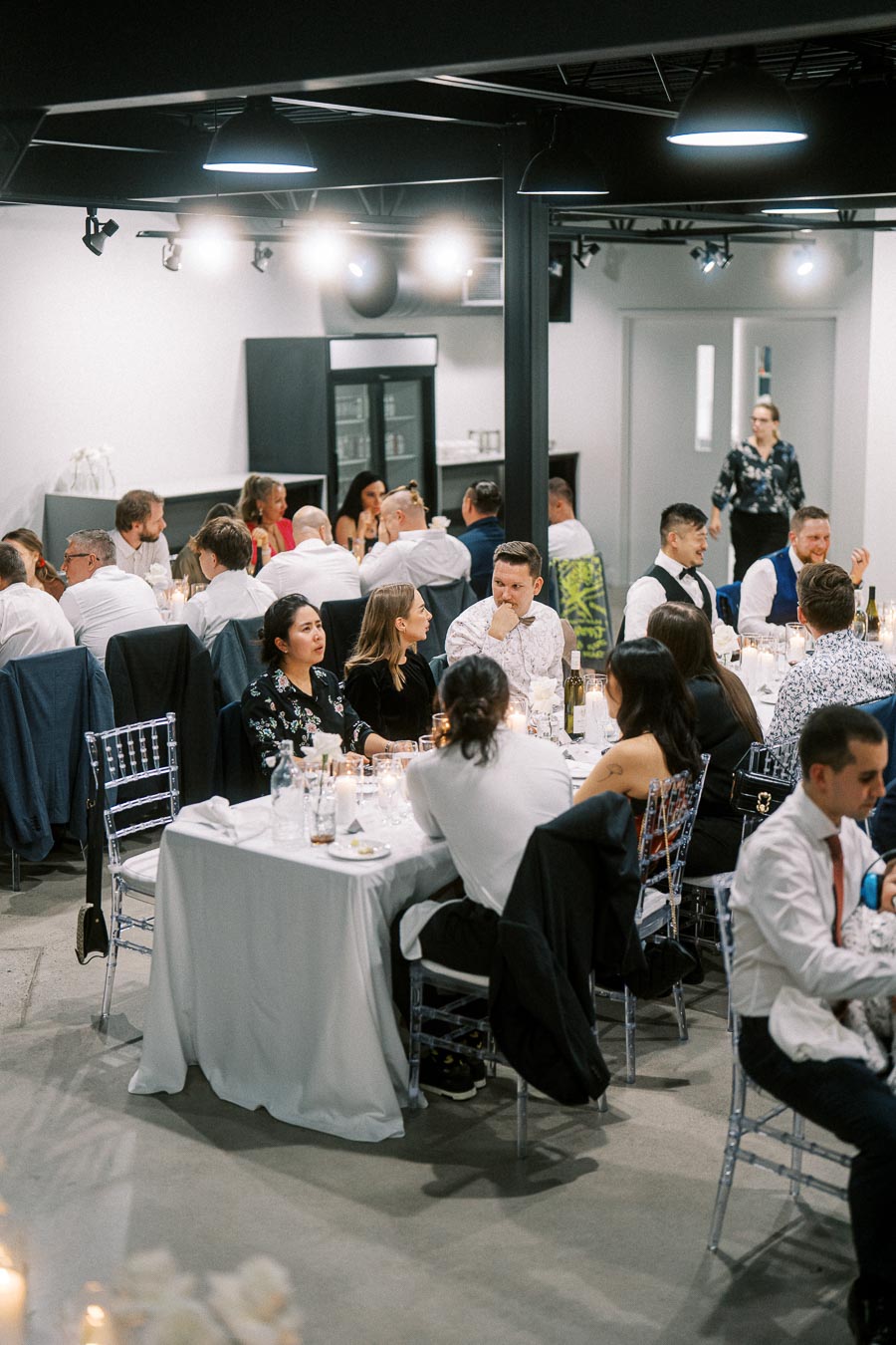 A lively gathering of people dressed in formal attire seated around tables in a modern event space, engaged in conversation during a social or corporate event.