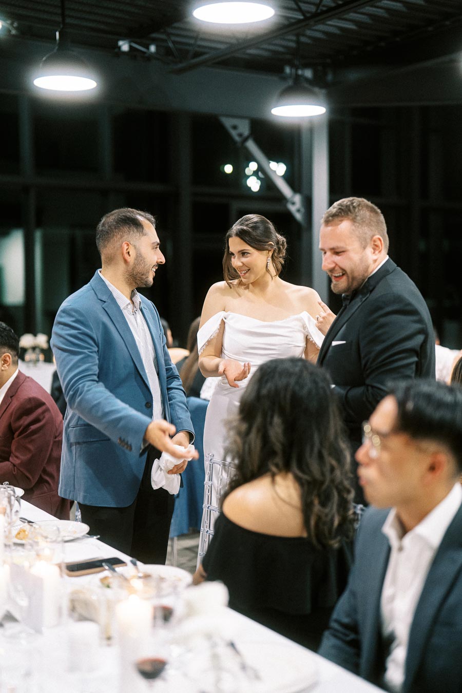 A group of well-dressed people enjoying a formal dinner event, with a woman in a white dress engaging in conversation with two men in suits under modern lighting.