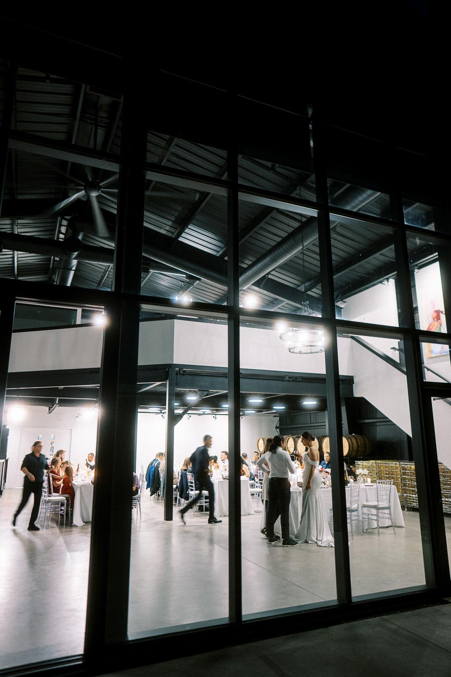 A lively wedding reception viewed through large windows at night, featuring elegantly dressed guests seated at tables inside a modern industrial venue with exposed beams and ambient lighting.