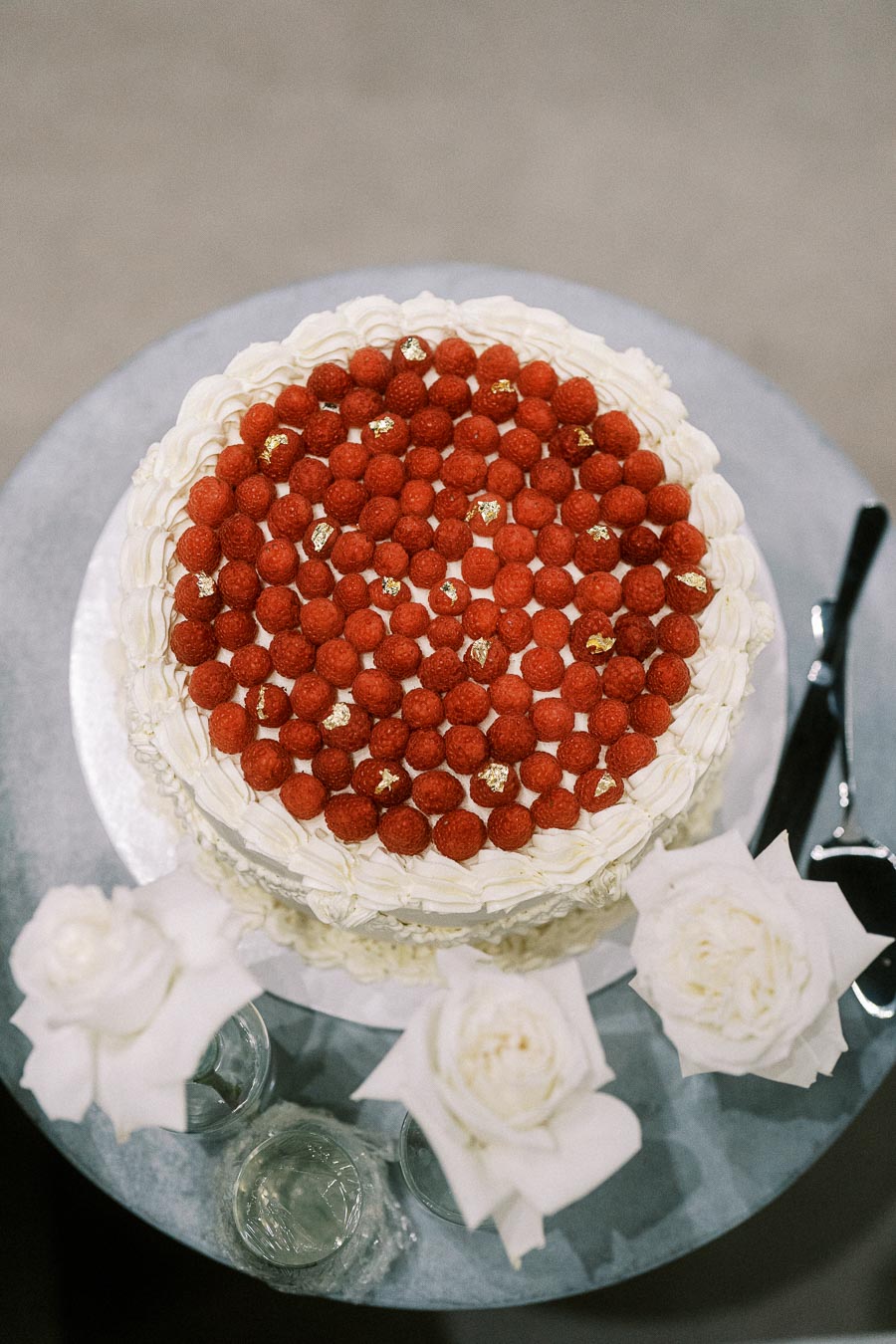 A beautifully decorated white frosted cake topped with fresh raspberries and gold leaf accents on a round table, accompanied by elegant white roses and a knife.