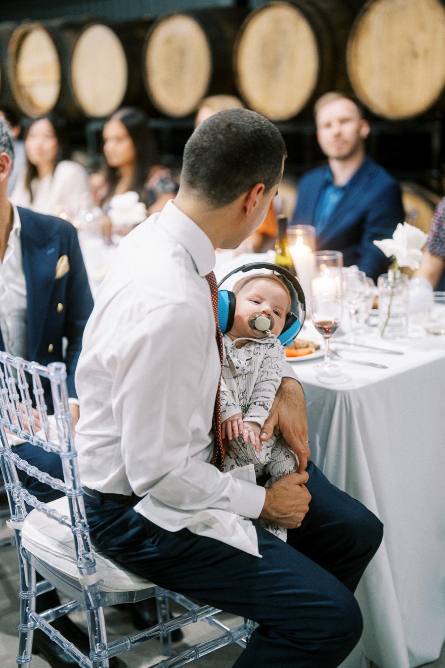 Father holding baby wearing noise-canceling headphones at a wedding reception, surrounded by guests and elegantly decorated tables with wine and candles.
