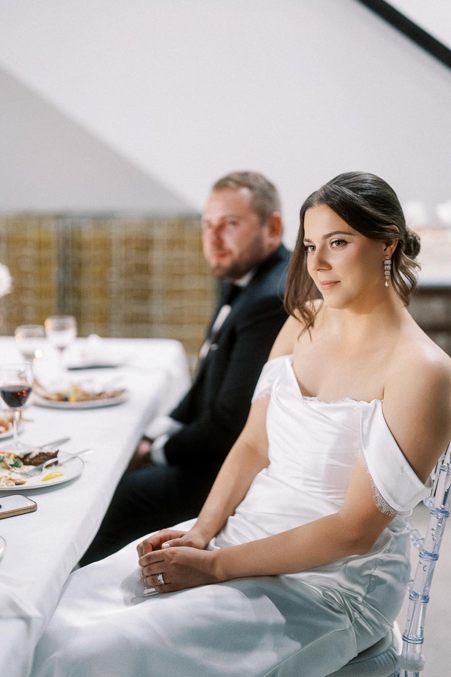 Bride sitting at a wedding reception table in a white dress, with a groom in a suit in the background; plates of food and glasses are visible on the table.