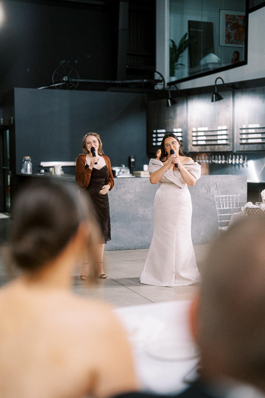 Two women holding microphones are singing at an indoor event, with blurred guests in the foreground, and a modern setting featuring sleek countertops and a contemporary design in the background.