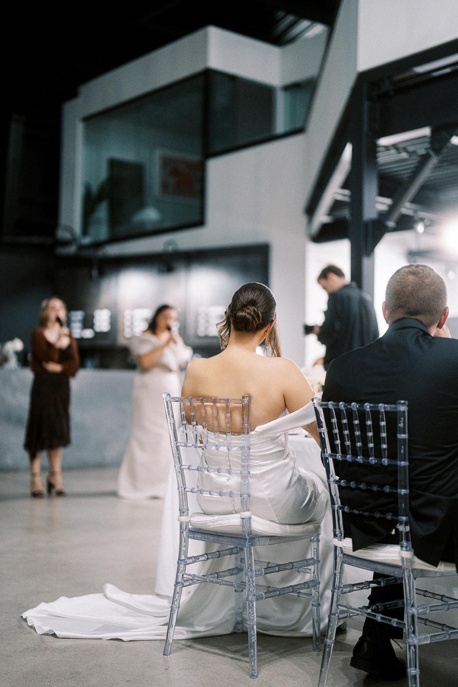 Wedding reception with bride and groom seated on transparent chairs, listening to speeches in a modern venue.