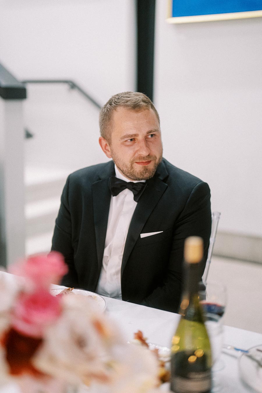 A man in a formal black suit and bow tie sitting at an elegantly set dining table with flowers and wine, in a modern and stylish indoor setting.