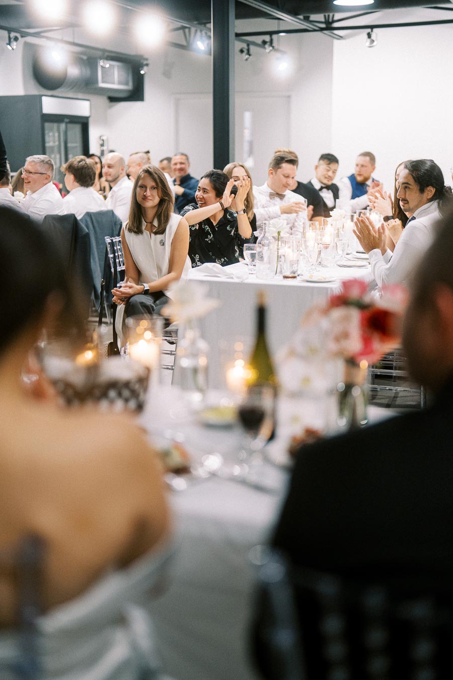 A group of elegantly dressed people seated at a wedding reception, with tables adorned with candles and flowers, creating a festive and joyful atmosphere indoors.