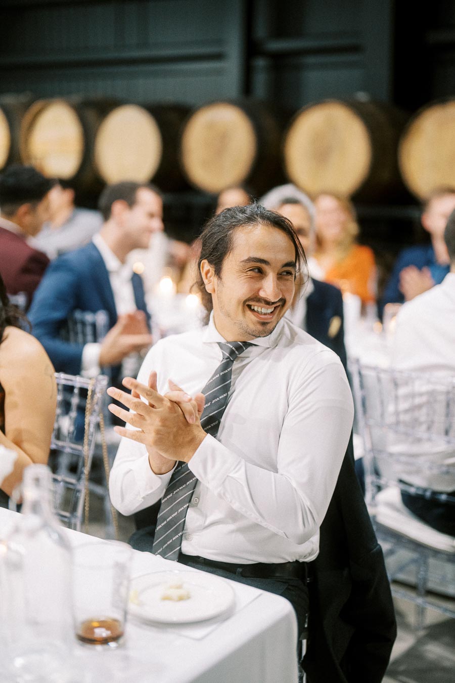 Smiling man in a formal setting clapping, surrounded by people at a dinner event with wooden barrels in the background.