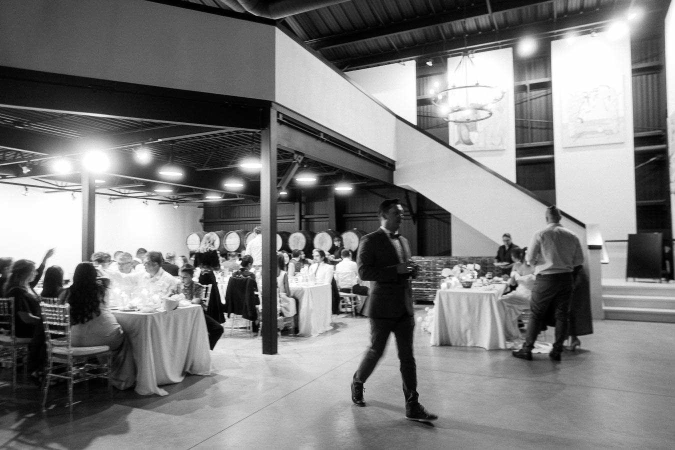 Black and white image of a formal event inside a modern industrial venue, featuring elegantly dressed guests seated at tables adorned with tablecloths, with wine barrels in the background.
