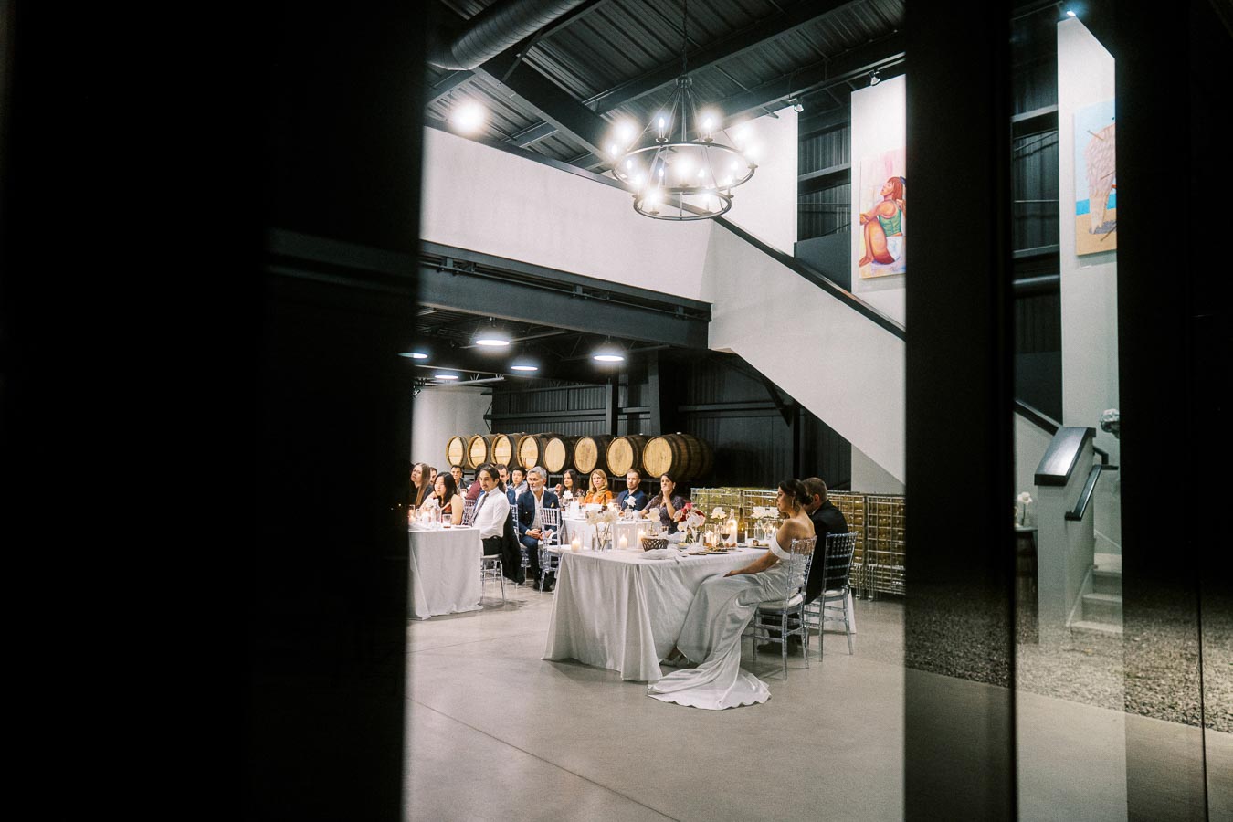Elegant wedding reception inside a modern wine cellar, featuring guests seated at decorated tables under ambient lighting with wooden barrels and contemporary art in the background.