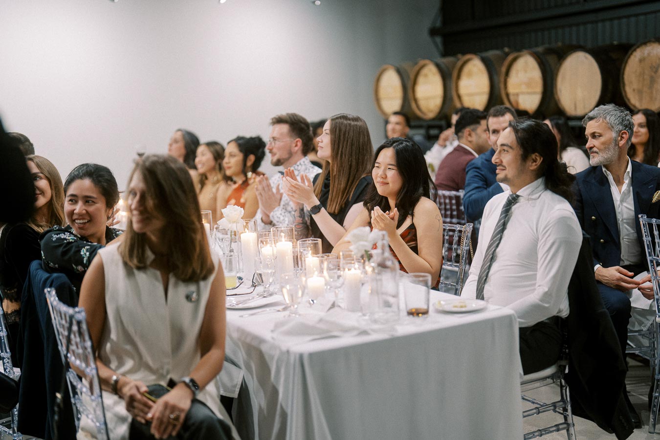 A group of people seated at elegantly decorated tables with lit candles in a cozy event space, enjoying a gathering with a backdrop of wooden barrels.