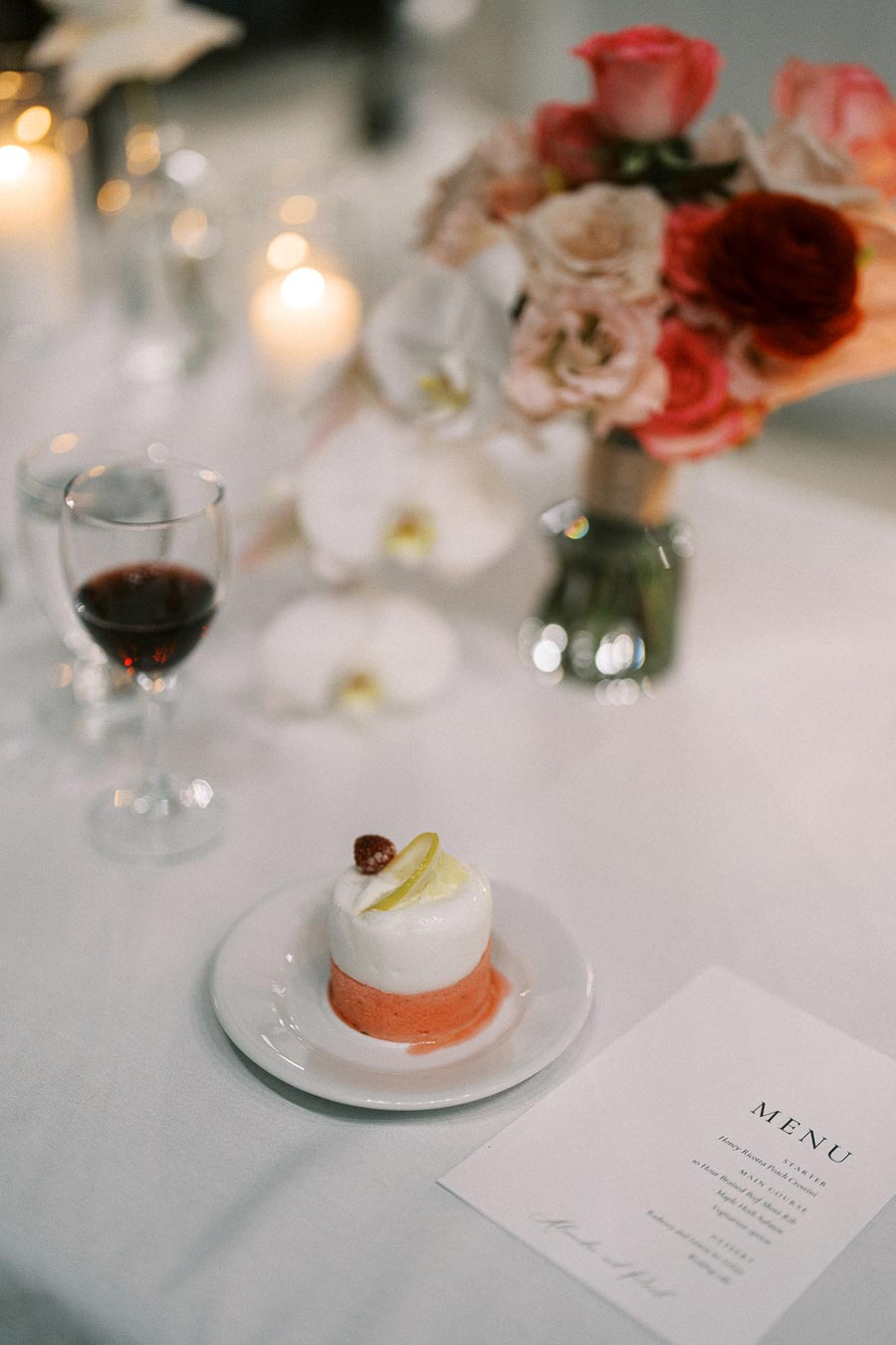 A romantic table setting with a delicate dessert featuring layers of white and pink mousse garnished with a raspberry and lemon slice on a white plate. Nearby, a glass of red wine, a floral centerpiece with pink, red, and white flowers, and a menu card complete the elegant ambiance.