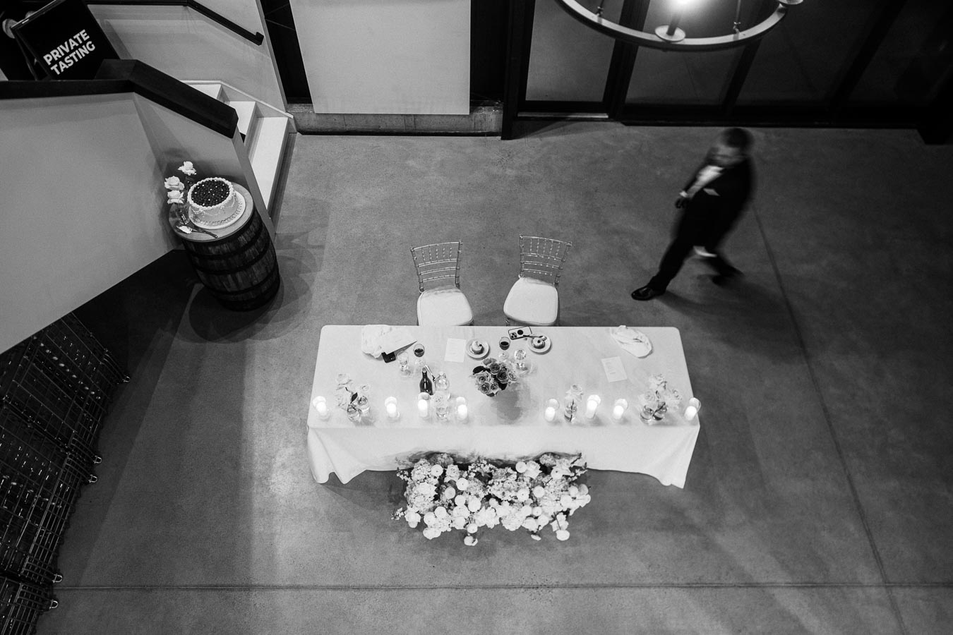 Overhead view of an elegantly set table for a private tasting, featuring candles and floral arrangements, with a person walking nearby, captured in black and white.