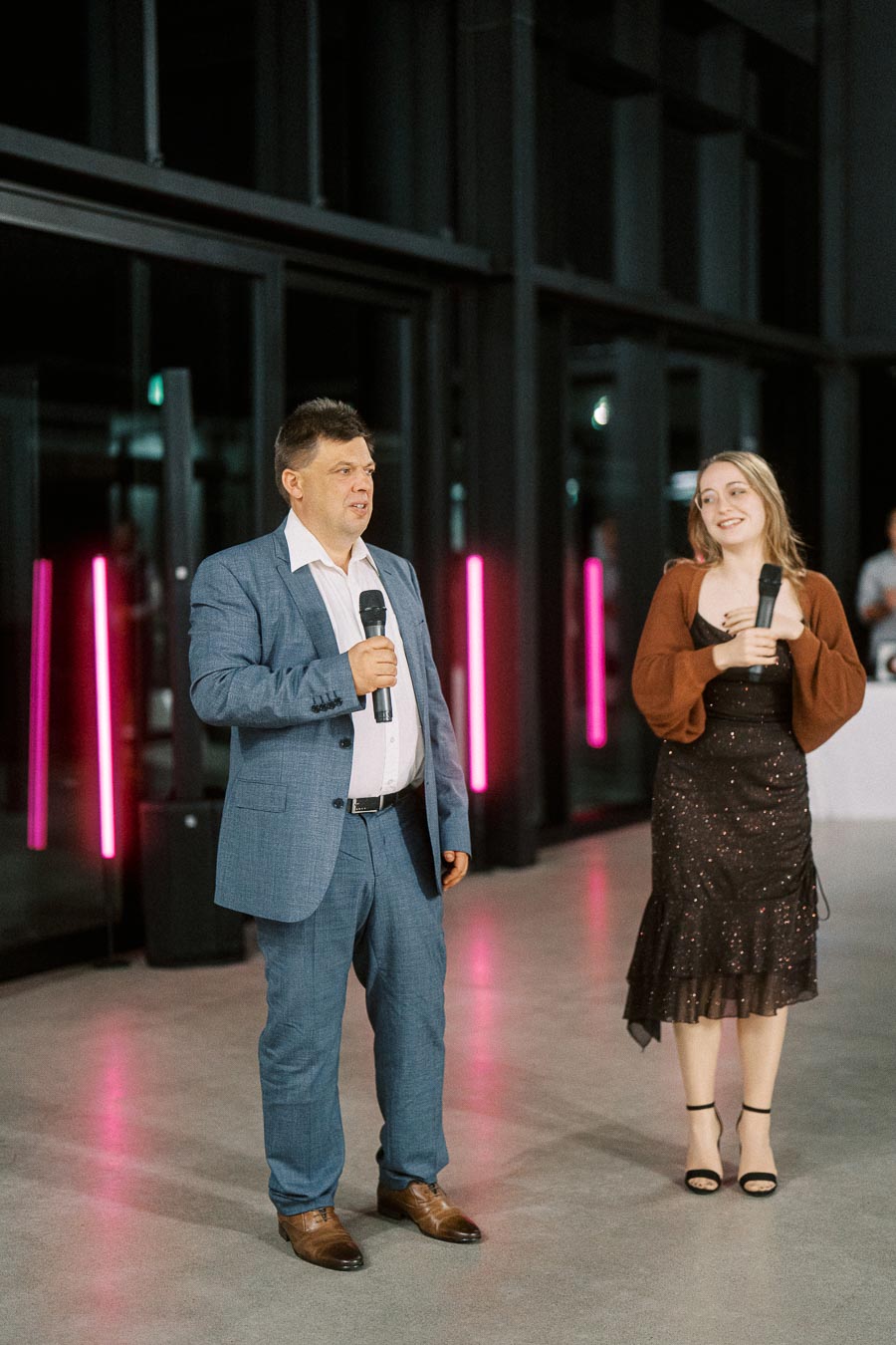 Two people holding microphones and speaking at an indoor event, both dressed formally with neon lights in the background.